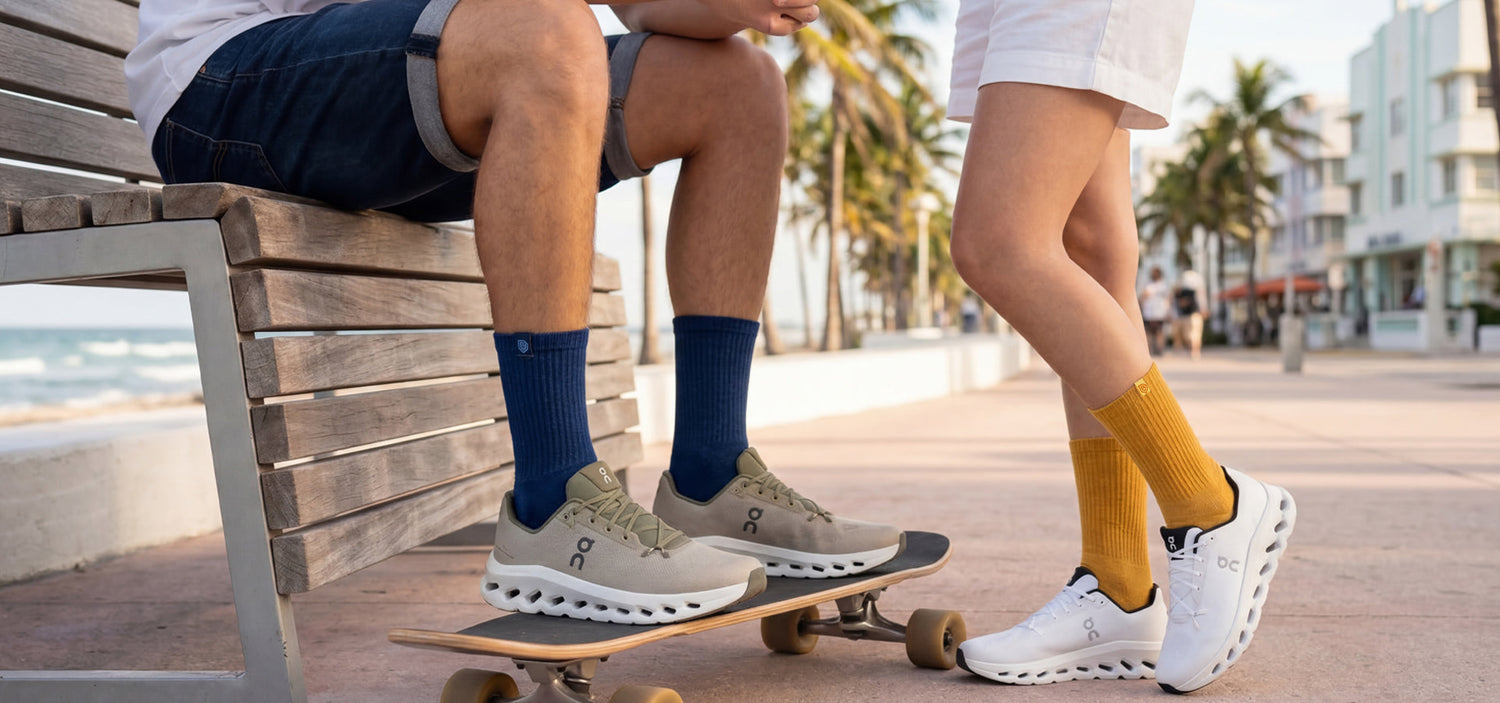 Two friends in Bulg crew socks relaxing on a beach promenade bench with palm trees and skateboard